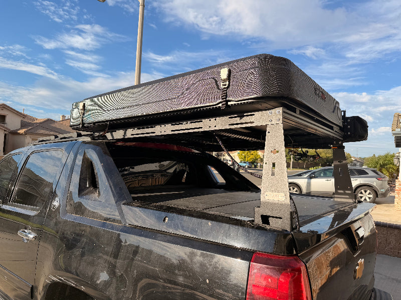 A black hardtop truck roof box mounted on a metal rack system installed on the bed of a dark pickup truck, photographed outdoors under a blue sky with clouds.
